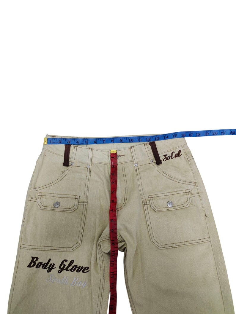 Puede incluir: Pantalones vaqueros de color caqui con costuras y detalles marrones. Cuenta con dos bolsillos delanteros, cierre de bot&oacute;n y el texto "Body Glove South Bay" bordado en la parte delantera. La palabra "SoCal" est&aacute; bordada en la parte trasera. Una cinta m&eacute;trica est&aacute; colocada sobre los pantalones.