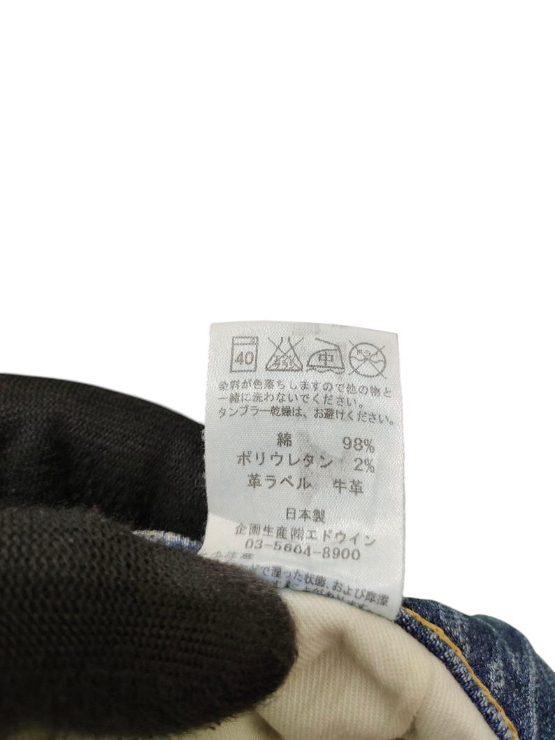 May include: A close-up of a clothing tag with Japanese text and care symbols. The tag lists the materials as 98% cotton and 2% polyurethane. The tag also includes the text "Made in Japan".