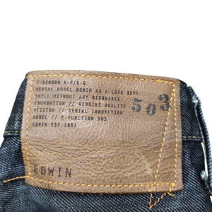 Op de afbeelding: Close-up van de achterkant van een donkerblauwe denim jeans. Een bruin leren label is op de jeans genaaid, met het merk "EDWIN" en het nummer "503". Het label bevat ook tekst over de kwaliteit van de denim en de oprichting van het merk in 1951.