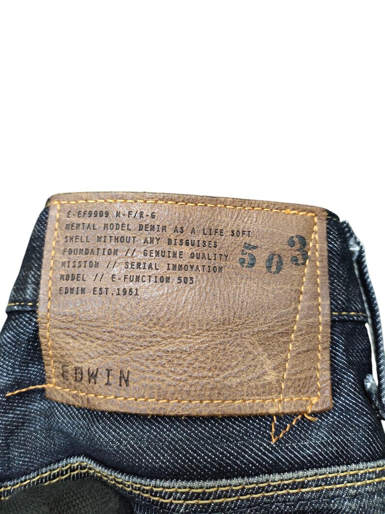 Op de afbeelding: Close-up van de achterkant van een donkerblauwe denim jeans. Een bruin leren label is op de jeans genaaid, met het merk "EDWIN" en het nummer "503". Het label bevat ook tekst over de kwaliteit van de denim en de oprichting van het merk in 1951.