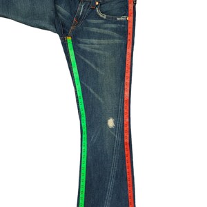 K&ouml;nnte beinhalten: Eine blaue Jeanshose mit Bootcut-Bein. Die Jeans hat eine helle Waschung und ist mit einem kleinen Loch am linken Bein abgenutzt. Die Jeans wird mit einem gr&uuml;nen und roten Ma&szlig;band gemessen.