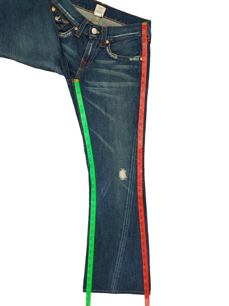 K&ouml;nnte beinhalten: Eine blaue Jeanshose mit Bootcut-Bein. Die Jeans hat eine helle Waschung und ist mit einem kleinen Loch am linken Bein abgenutzt. Die Jeans wird mit einem gr&uuml;nen und roten Ma&szlig;band gemessen.