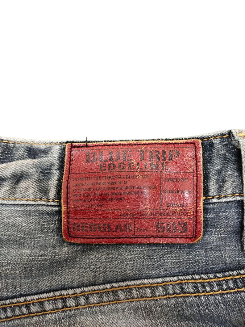 May include: Close-up of the back of blue denim jeans. A red leather patch with the text "BLUE TRIP EDGELINE" is visible. The jeans have a regular fit and are stitched with yellow thread.