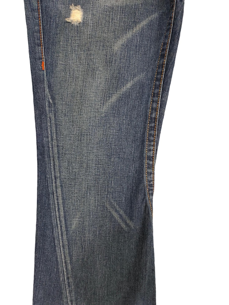 K&ouml;nnte beinhalten: Nahaufnahme des Jeansstoffes einer blauen Jeans mit einem kleinen Loch im Stoff. Die Jeans hat orangefarbene N&auml;hte entlang der N&auml;hte.