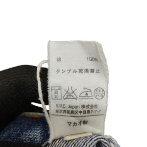 Puede incluir: Primer plano de una etiqueta de ropa blanca con texto japon&eacute;s y s&iacute;mbolos de cuidado. La etiqueta indica "100%" e instrucciones de lavado. Tambi&eacute;n se ve la marca "A.P.C. Japan".
