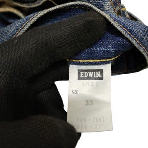 Puede incluir: Primer plano de unos vaqueros de mezclilla azules. La etiqueta indica "EDWIN", el modelo "503Z" y la talla "33". Los vaqueros est&aacute;n desgastados y muestran signos de uso.