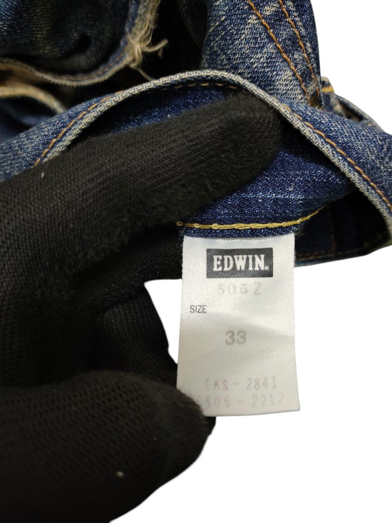 Puede incluir: Primer plano de unos vaqueros de mezclilla azules. La etiqueta indica "EDWIN", el modelo "503Z" y la talla "33". Los vaqueros est&aacute;n desgastados y muestran signos de uso.