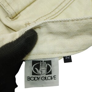 Puede incluir: Primer plano de tejido vaquero beige claro. La imagen muestra detalles de costura, una etiqueta con el logo "BODY GLOVE" y una etiqueta de talla 32. La tela parece ser de un par de vaqueros o prenda similar.