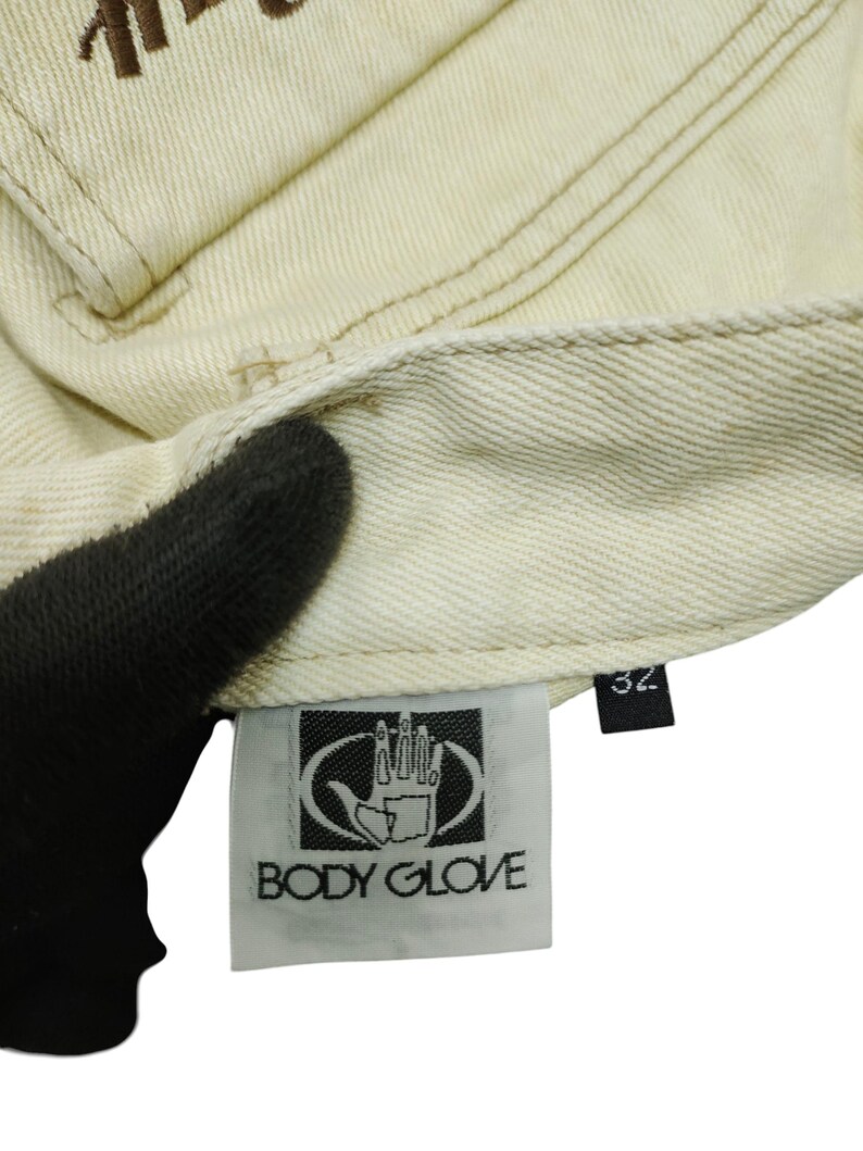 Puede incluir: Primer plano de tejido vaquero beige claro. La imagen muestra detalles de costura, una etiqueta con el logo "BODY GLOVE" y una etiqueta de talla 32. La tela parece ser de un par de vaqueros o prenda similar.