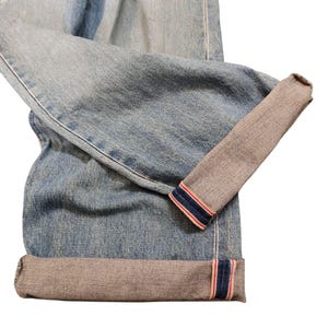 May include: Light blue denim jeans with cuffed hems. The cuffs reveal a contrasting gray fabric with red and blue stripes. The jeans are folded, showcasing the texture and color variations of the denim.