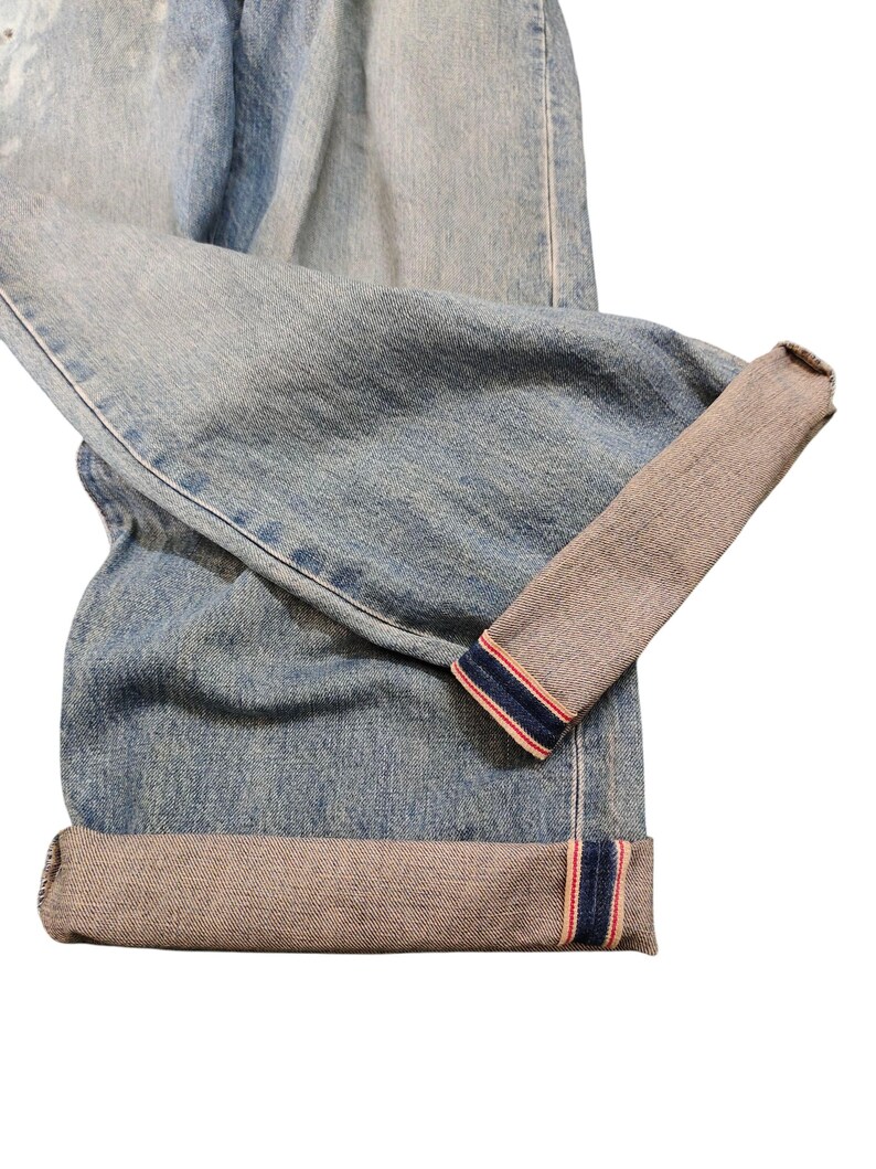 May include: Light blue denim jeans with cuffed hems. The cuffs reveal a contrasting gray fabric with red and blue stripes. The jeans are folded, showcasing the texture and color variations of the denim.