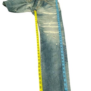 May include: Light blue denim jeans with distressed details, measuring approximately 40 inches in length. The jeans have a classic five-pocket design and are measured with a yellow and blue measuring tape.
