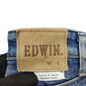 May include: Close-up of the back of blue denim jeans. A brown leather patch with the brand name "EDWIN" is stitched onto the jeans. Below the patch is a white tag that reads "Tokyo Japan Quality Jeans."