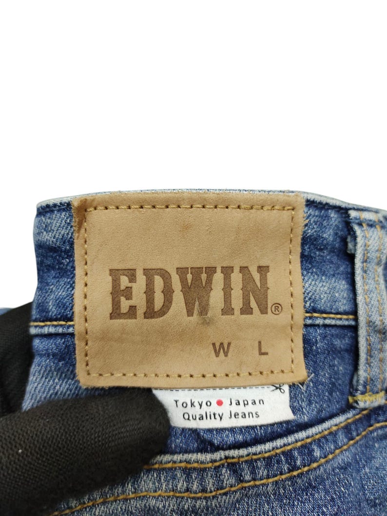 May include: Close-up of the back of blue denim jeans. A brown leather patch with the brand name "EDWIN" is stitched onto the jeans. Below the patch is a white tag that reads "Tokyo Japan Quality Jeans."