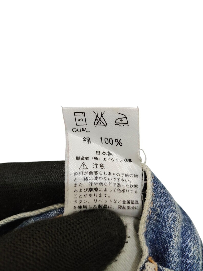 Puede incluir: Una etiqueta blanca con texto y s&iacute;mbolos japoneses, adherida a una tela vaquera. La etiqueta incluye instrucciones de lavado y el texto "綿 100%" que indica 100% algod&oacute;n. La etiqueta tambi&eacute;n tiene el texto "日本製" que significa "Hecho en Jap&oacute;n".