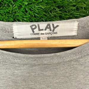 Może przedstawiać: Szara koszulka z czarnym nadrukiem graficznym słowa "PLAY" i tekstem "COMME des GAR&Ccedil;ONS" na białej metce.