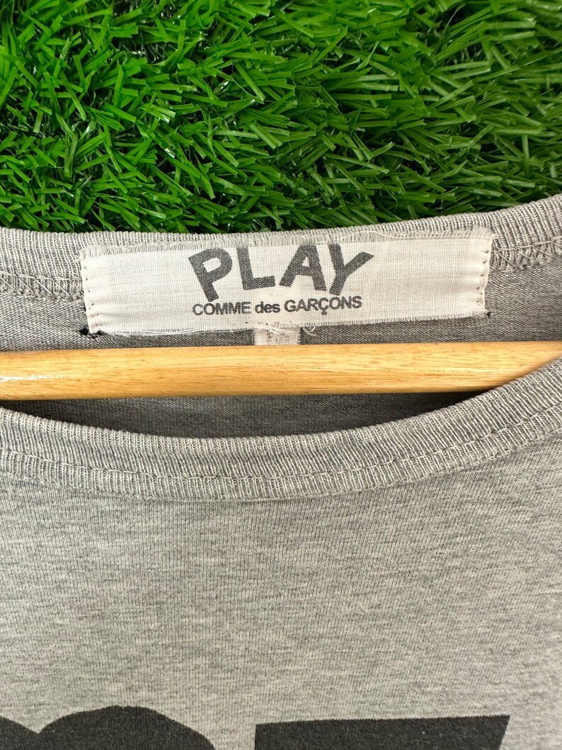 Może przedstawiać: Szara koszulka z czarnym nadrukiem graficznym słowa "PLAY" i tekstem "COMME des GAR&Ccedil;ONS" na białej metce.