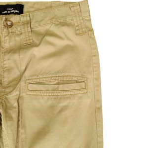 Puede incluir: Un pantal&oacute;n cargo beige con una etiqueta negra que dice "tricot COMME des GARCONS" en la cintura.