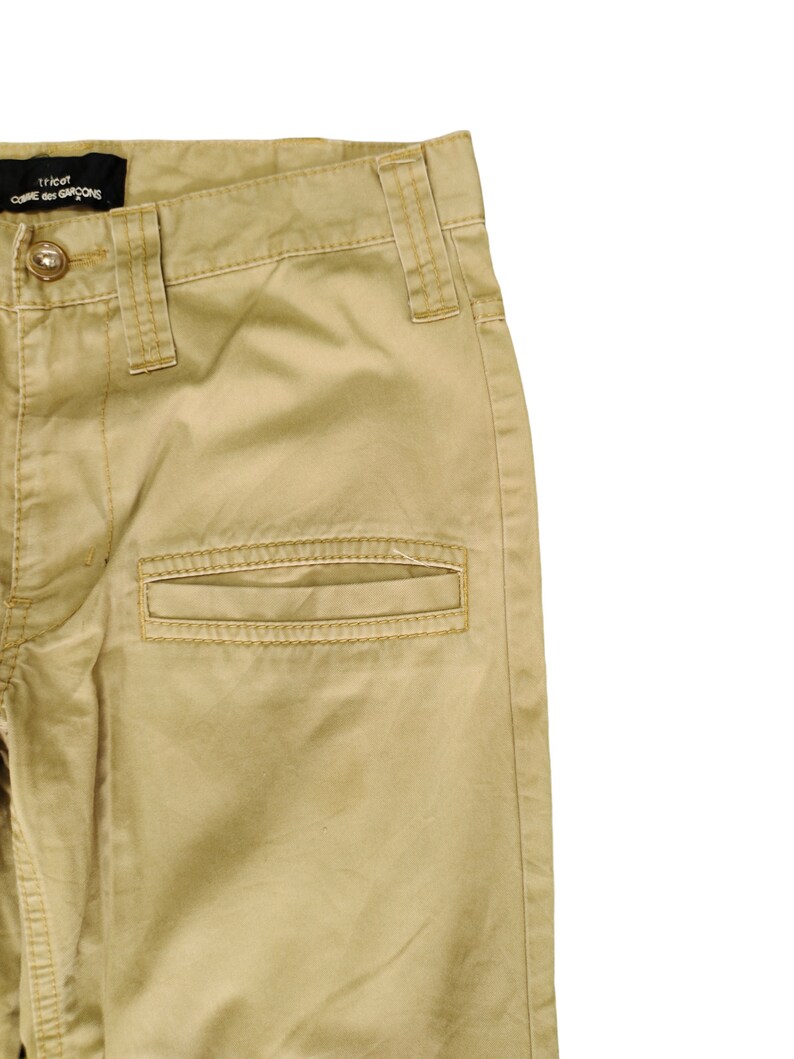 Puede incluir: Un pantal&oacute;n cargo beige con una etiqueta negra que dice "tricot COMME des GARCONS" en la cintura.