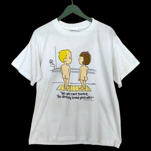 May include: White t-shirt featuring a cartoon illustration of a boy and girl standing in a bath. The text on the shirt reads "No you can't touch it. You already broke yours off!!!"