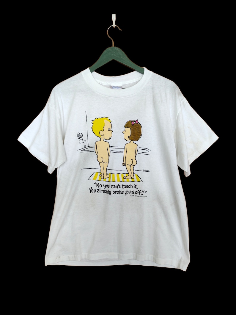 May include: White t-shirt featuring a cartoon illustration of a boy and girl standing in a bath. The text on the shirt reads "No you can't touch it. You already broke yours off!!!"