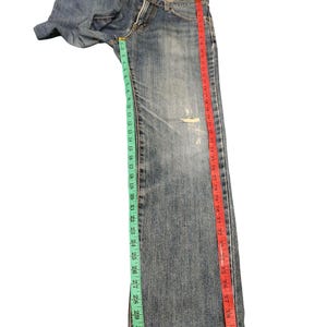 May include: Blue denim jeans with a distressed look, featuring a rip on the leg. The jeans are measured with a green and red measuring tape. The inseam measures approximately 32 inches.