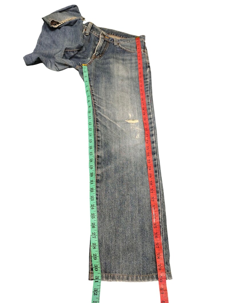 May include: Blue denim jeans with a distressed look, featuring a rip on the leg. The jeans are measured with a green and red measuring tape. The inseam measures approximately 32 inches.