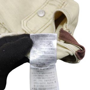 Puede incluir: Primer plano de una chaqueta beige con cierre de bot&oacute;n y un acento marr&oacute;n. Una etiqueta con texto japon&eacute;s es visible. La chaqueta parece estar hecha de una tela duradera.