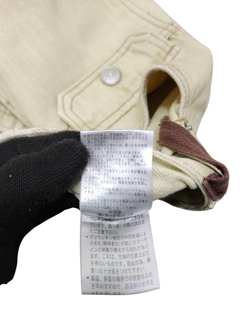Puede incluir: Primer plano de una chaqueta beige con cierre de bot&oacute;n y un acento marr&oacute;n. Una etiqueta con texto japon&eacute;s es visible. La chaqueta parece estar hecha de una tela duradera.