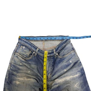 May include: Blue denim jeans with distressed detailing. A yellow measuring tape shows a waist measurement of approximately 18 inches. The jeans have a button fly and classic five-pocket design. The jeans are laid flat on a white surface.