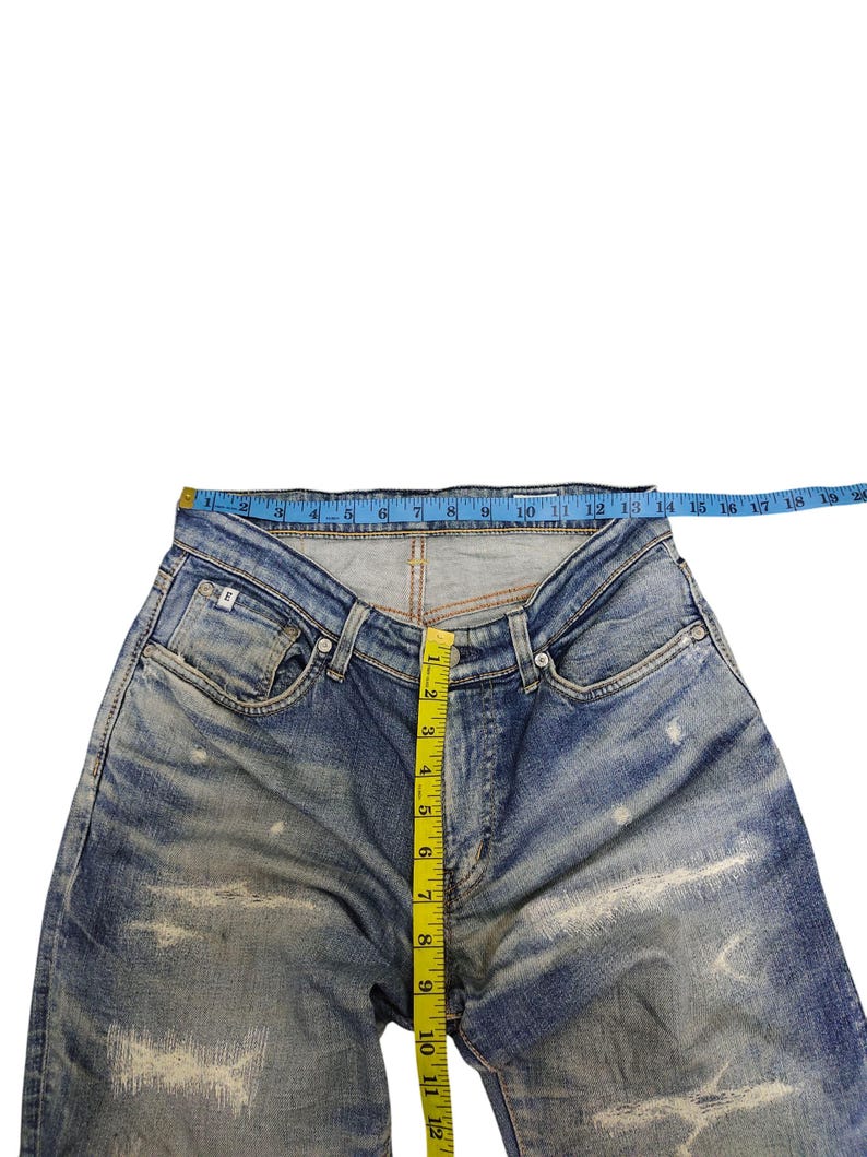 May include: Blue denim jeans with distressed detailing. A yellow measuring tape shows a waist measurement of approximately 18 inches. The jeans have a button fly and classic five-pocket design. The jeans are laid flat on a white surface.