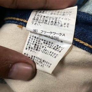 以下が含まれることがあります： 青いデニム衣類に付いた、日本語のテキストが書かれた白いタグのクローズアップ。タグには会社のロゴと、洗濯に関する注意書きなどのケア方法が記載されています。