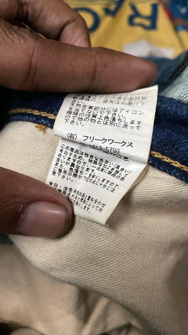 以下が含まれることがあります： 青いデニム衣類に付いた、日本語のテキストが書かれた白いタグのクローズアップ。タグには会社のロゴと、洗濯に関する注意書きなどのケア方法が記載されています。