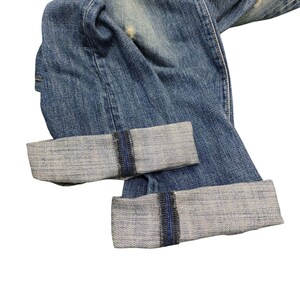 Puede incluir: Primer plano de unos vaqueros de mezclilla azules con los pu&ntilde;os doblados. Los vaqueros tienen un lavado descolorido y detalles desgastados. Los pu&ntilde;os est&aacute;n doblados, revelando la mezclilla de color m&aacute;s claro en el interior. Estilo cl&aacute;sico.