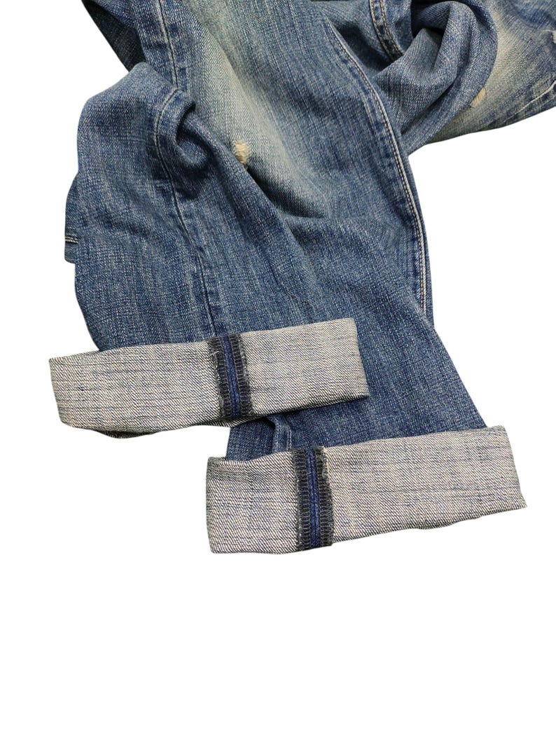 Puede incluir: Primer plano de unos vaqueros de mezclilla azules con los pu&ntilde;os doblados. Los vaqueros tienen un lavado descolorido y detalles desgastados. Los pu&ntilde;os est&aacute;n doblados, revelando la mezclilla de color m&aacute;s claro en el interior. Estilo cl&aacute;sico.