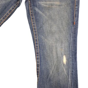 May include: Close-up of the bottom of a pair of blue denim jeans with a small faded spot. The jeans have orange stitching along the seams.