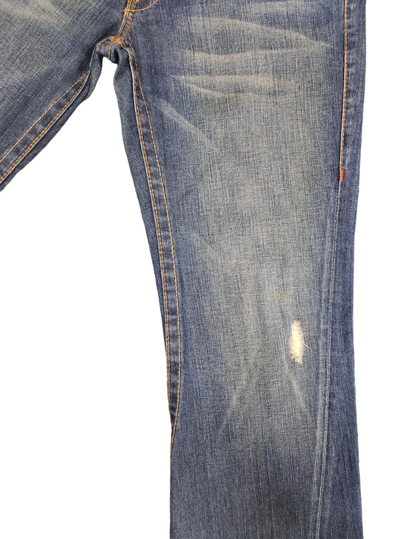 May include: Close-up of the bottom of a pair of blue denim jeans with a small faded spot. The jeans have orange stitching along the seams.