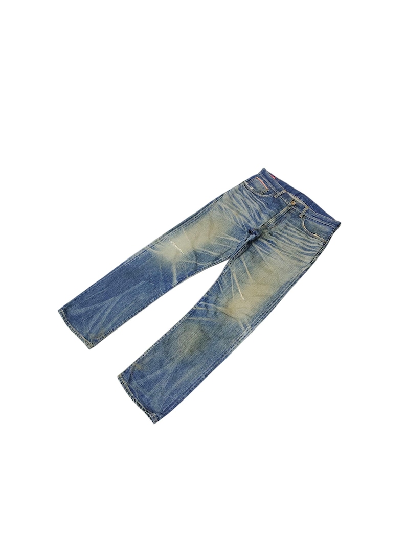 Puede incluir: Vaqueros de mezclilla azul descolorido con un dise&ntilde;o cl&aacute;sico de cinco bolsillos. Los jeans muestran signos de desgaste con ligeros desgastes y decoloraci&oacute;n en toda la tela. Los jeans est&aacute;n planos sobre un fondo blanco.