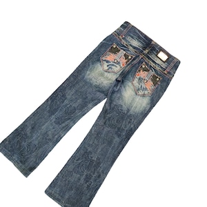May include: Blue denim jeans with a faded wash and decorative floral pattern. The back pockets feature embroidered designs with red and black accents. The jeans have a bootcut leg opening.