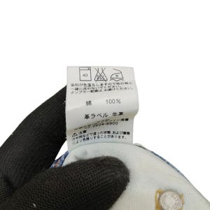 May include: Close-up of a clothing label with Japanese text and care symbols. The label is white with black text, indicating washing instructions and material composition. The fabric is 100% cotton. A hand wearing a black glove holds the label.