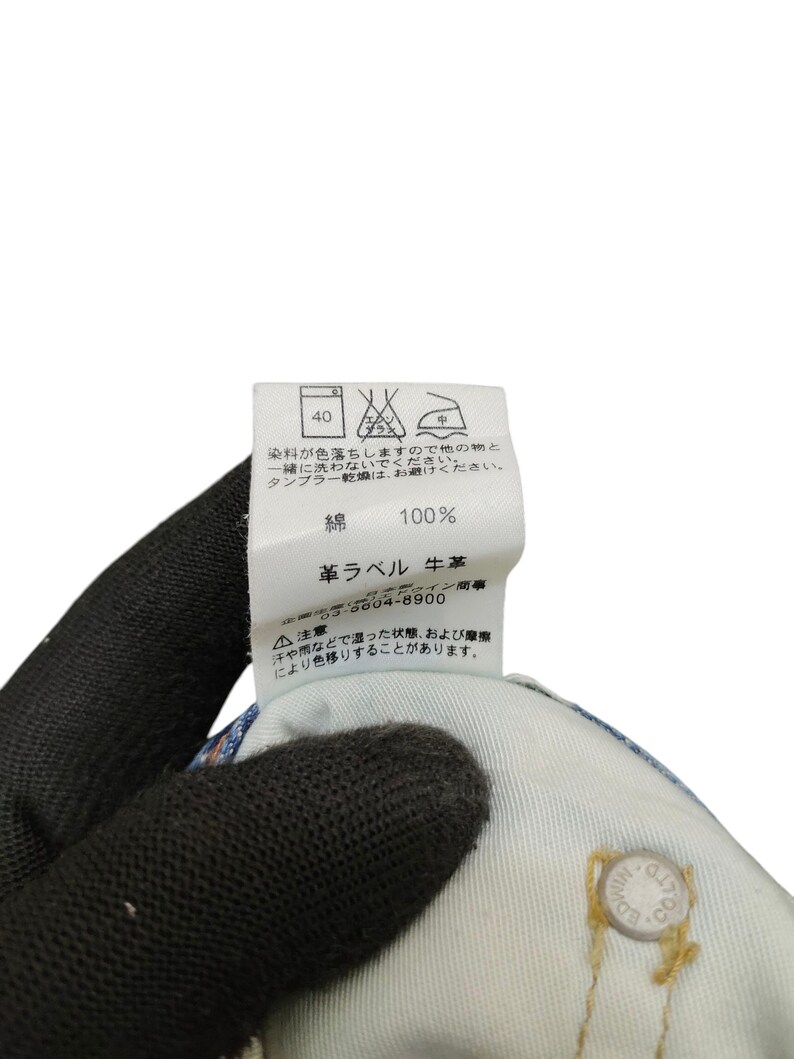 May include: Close-up of a clothing label with Japanese text and care symbols. The label is white with black text, indicating washing instructions and material composition. The fabric is 100% cotton. A hand wearing a black glove holds the label.