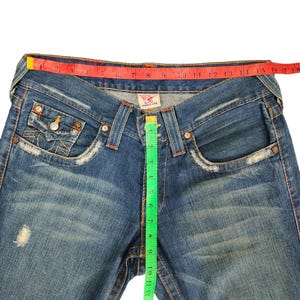 May include: Blue denim jeans with a red and white measuring tape around the waist and a green measuring tape down the side seam. The jeans have a distressed look with faded areas and small holes.
