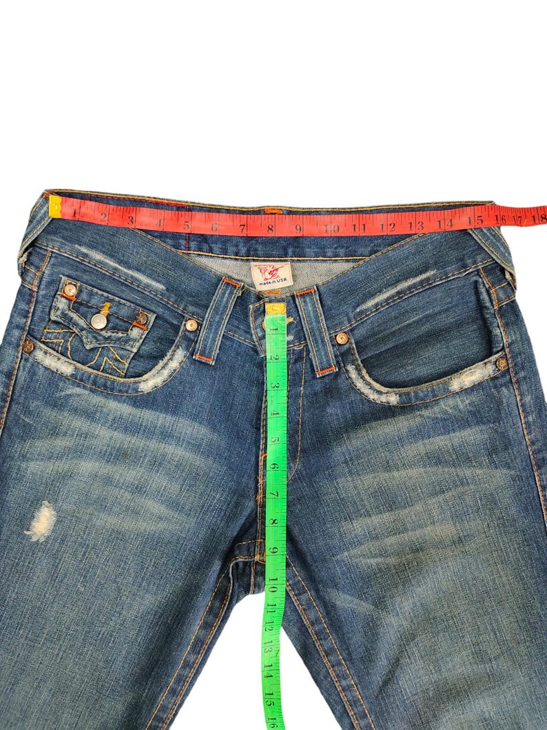 May include: Blue denim jeans with a red and white measuring tape around the waist and a green measuring tape down the side seam. The jeans have a distressed look with faded areas and small holes.