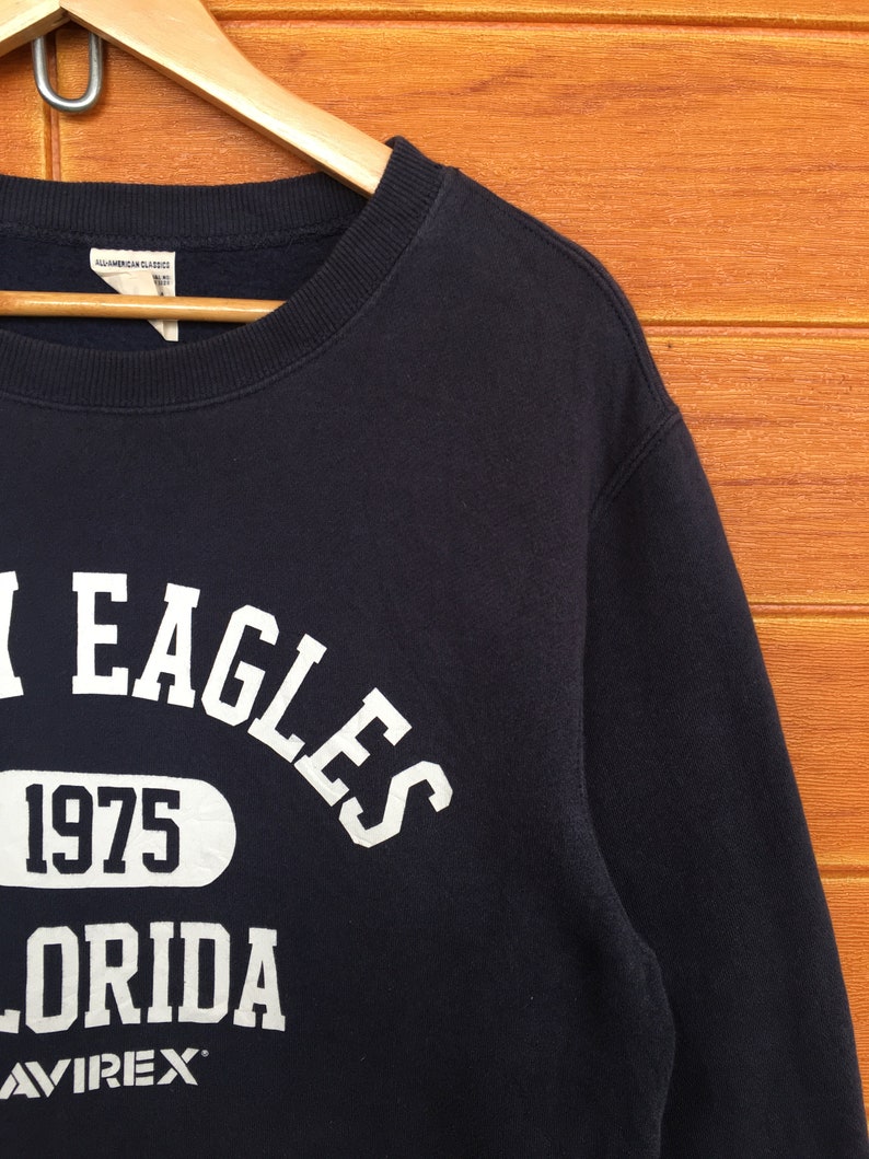 May include: A black sweatshirt with white text that reads "EAGLES 1975 FLORIDA AVIREX".