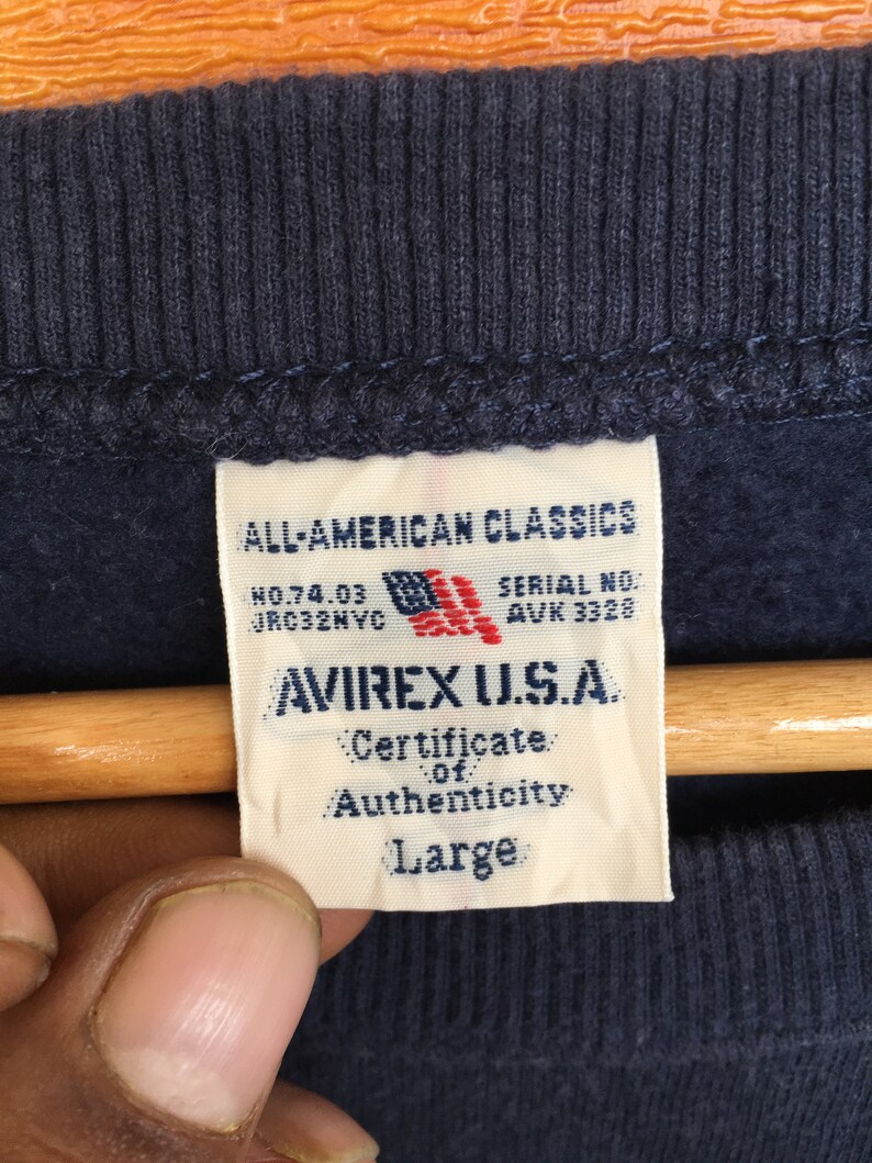 May include: A navy blue sweatshirt with a white tag that reads "ALL-AMERICAN CLASSICS", "NO.74.03 JRC32NVC", "SERIAL NO. AUK 3328", "AVIREX U.S.A.", "Certificate of Authenticity", and "Large".