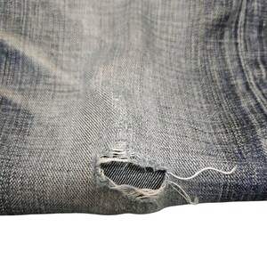 May include: Close-up of distressed denim fabric. The light blue jeans have a worn appearance with a visible tear, revealing the inner weave. Frayed threads extend from the ripped area, adding to the vintage aesthetic.