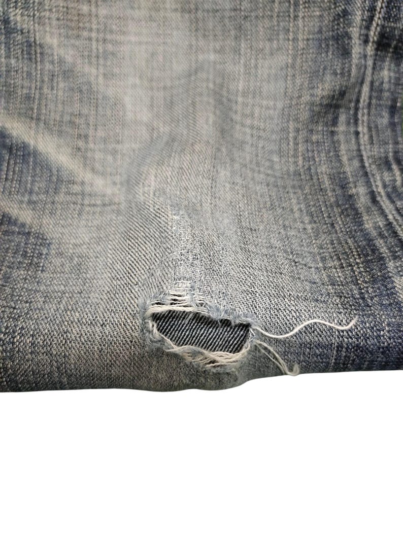 May include: Close-up of distressed denim fabric. The light blue jeans have a worn appearance with a visible tear, revealing the inner weave. Frayed threads extend from the ripped area, adding to the vintage aesthetic.