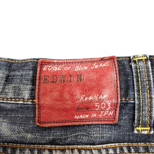 May include: Close-up of blue denim jeans with a red leather patch. The patch reads "EDGE OF BLUE JEANS" and "EDWIN" in white lettering. The jeans have yellow stitching and a "Regular 503" label. Made in JPN.