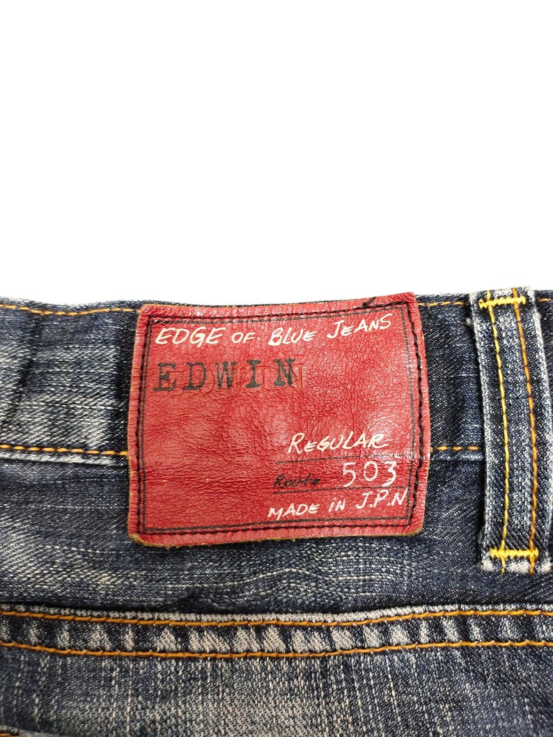 May include: Close-up of blue denim jeans with a red leather patch. The patch reads "EDGE OF BLUE JEANS" and "EDWIN" in white lettering. The jeans have yellow stitching and a "Regular 503" label. Made in JPN.