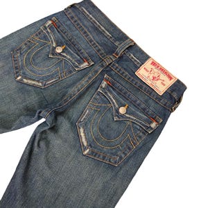 May include: A pair of blue denim jeans with a distressed look. The back pockets have a signature True Religion horseshoe design with stitching in orange and yellow thread. The jeans have a red leather patch with the True Religion logo and the words "True Religion Brand Jeans" printed on it.